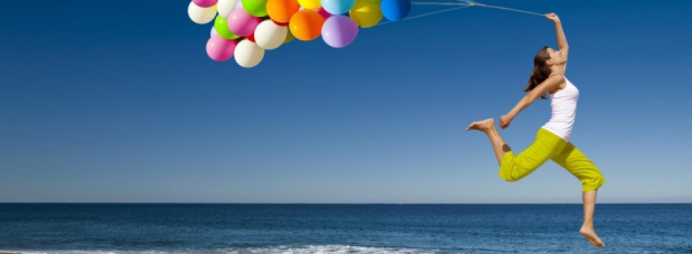 Nell’immagine una ragazza sospesa tra il mare e il cielo azzurro che tiene in mano dei palloncini colorati.