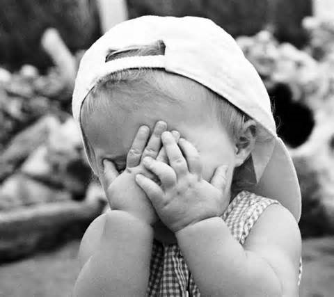 Foto in bianco e nero di un bimbo con un cappellino con visiera all'indietro che si copre la faccina con le mani