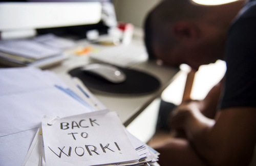 Immagine dove sullo sfondo sfocato si vede una persona con la testa china sulla scrivania che sta dormendo davanti ad un computer e in primo piano un post-it con la scritta BACK TO WORK!